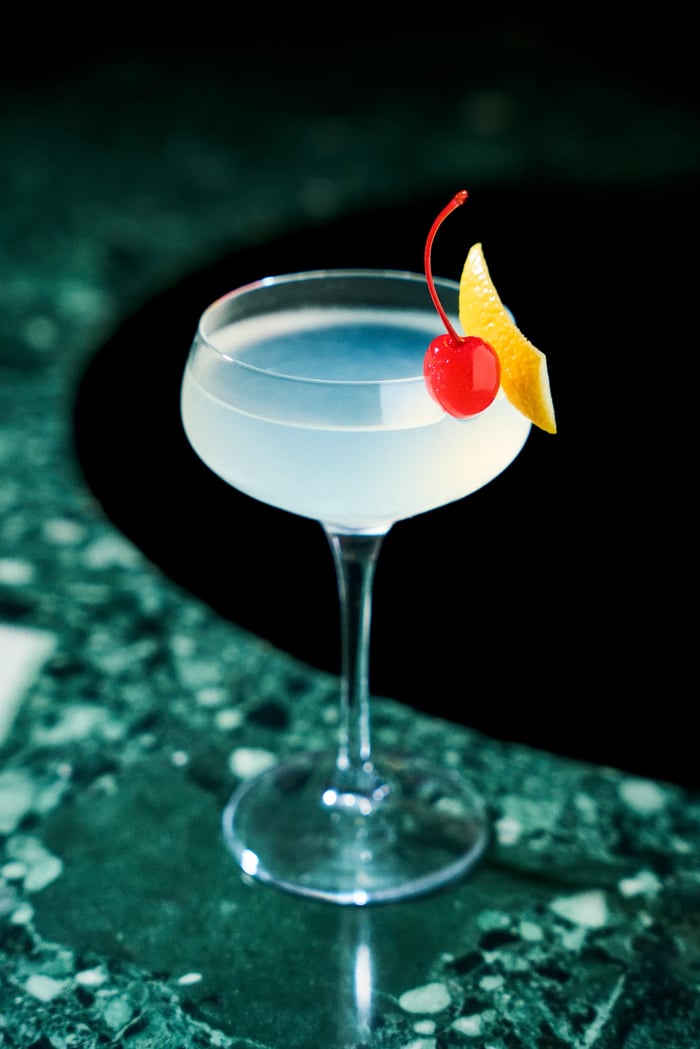 Cocktail bianco con ciliegia maraschino e scorza d'arancia in coppa coupe su marmo verde — drink photography Alchimia Milano