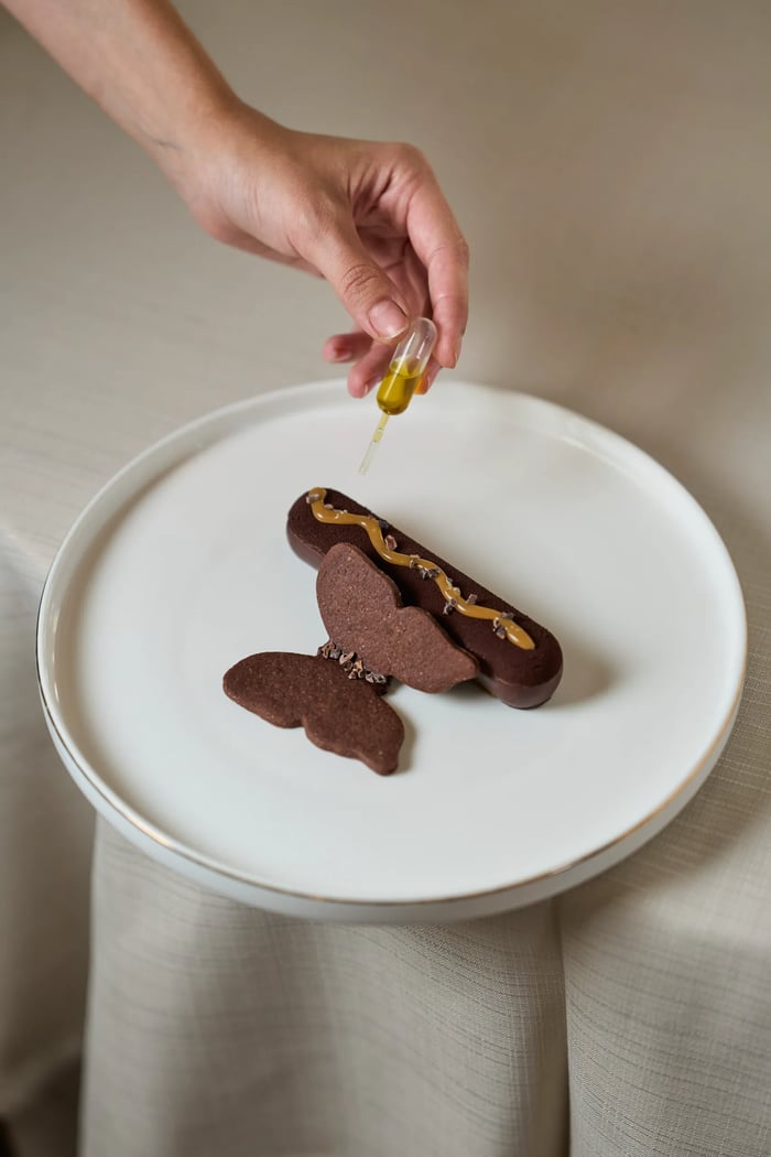 Dessert al cioccolato a forma di farfalla con caramello e granella di cacao, mano con pipetta d'olio — fotografia pasticceria gourmet Milano